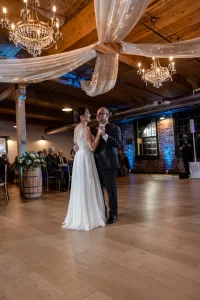 Professional Photographer A bride in a white dress dances with an older man, likely her father, in a warmly lit reception hall—an intimate moment captured through wedding photography. Chandeliers, draped fabric, wood floor, and exposed brick add to the scene as guests look on. www.maranonimagery.com