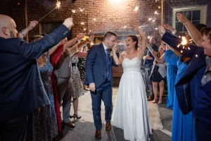 Professional Photographer A bride and groom walk hand in hand, smiling and laughing, while wedding guests holding sparklers celebrate around them at night—a magical moment of wedding photography set against a festive brick wall background. www.maranonimagery.com