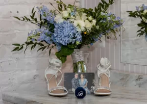 Professional Photographer A bouquet of blue and white flowers in a vase sits behind white high-heeled shoes, a photo of a couple, and a blue ring box with a B on it—all beautifully arranged by a wedding photographer on a marble surface. www.maranonimagery.com