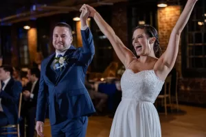 Professional Photographer A bride in a white dress and groom in a blue suit hold hands and smile joyfully during their wedding reception, surrounded by guests—capturing the perfect moment in timeless wedding photography within a warmly lit venue. www.maranonimagery.com
