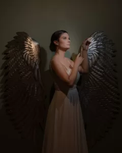 Professional Photographer A woman in a white dress stands in profile against a dark background, illuminated by soft light. Perfect for creative portraits, she has large metallic angel wings and gently touches her fingers together, gazing upward thoughtfully. www.maranonimagery.com