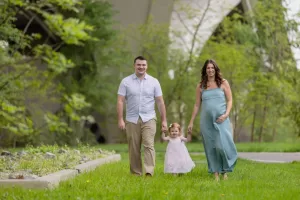 Professional Photographer A smiling family of three, captured by a photographer, including a pregnant woman in a blue dress, a man in a light shirt, and a young girl in pink, walk hand-in-hand on a grassy path outdoors. www.maranonimagery.com