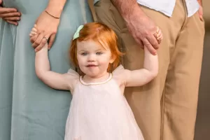 Professional Photographer A young red-haired girl in a white dress holds hands with two adults, one in a blue dress and the other in tan pants and a white shirt. Captured by a skilled photographer, the portrait shows the girl looking at the camera with a small smile. www.maranonimagery.com