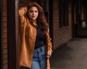 Professional Photographer A woman with long, wavy brown hair leans against a wooden post, wearing a tan corduroy jacket over a black top and blue jeans, standing on a pathway beside a wooden building—perfect for natural light portraits or headshots. www.maranonimagery.com