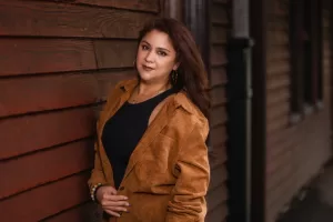 Professional Photographer A woman with long, wavy brown hair stands against a wooden wall, wearing a brown corduroy jacket over a black top. She looks at the camera with a neutral expression, her hand near her waist—an elegant portrait reminiscent of fine portraits photography. www.maranonimagery.com