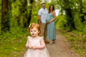 Professional Photographer A young red-haired girl in a light dress stands on a wooded path, captured by a talented photographer, with a man and pregnant woman blurred in the background among lush green trees. www.maranonimagery.com