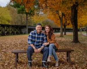Professional Photographer A smiling couple sits together on a wooden bench in a park during autumn, surrounded by fallen leaves and colorful trees, as a photographer captures their joy amidst the vibrant orange and yellow foliage. www.maranonimagery.com