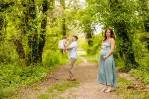 Professional Photographer A pregnant woman in a light blue dress stands on a forest path, smiling at the camera, while a man lifts a young girl in pink. This charming portrait captures memories amid lush green trees. www.maranonimagery.com