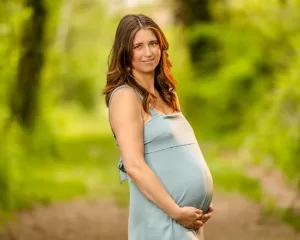 Professional Photographer A pregnant woman with long brown hair, wearing a light blue sleeveless dress, stands outdoors on a green, blurred forest path, gently holding her belly and smiling softly at the camera—perfect for captivating maternity portraits. www.maranonimagery.com