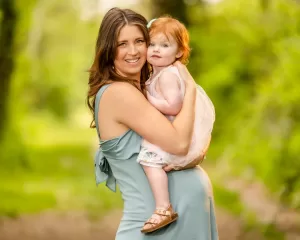 Professional Photographer A woman in a light blue dress holds a young girl with red hair and a white dress outdoors, surrounded by lush greenery on a sunlit path. Both are looking at the camera, smiling—capturing the magic of the moment with an expert photographer’s touch. www.maranonimagery.com
