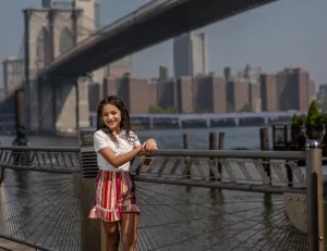 Professional Photographer A young girl stands smiling by a waterfront railing with the Brooklyn Bridge and city buildings in the background on a sunny day, perfectly capturing a headshot any photographer would cherish. www.maranonimagery.com