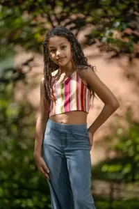 Professional Photographer A young girl with long, curly hair stands outdoors, posing confidently for a photographer in a colorful striped crop top and high-waisted blue jeans, with greenery and soft sunlight in the background. www.maranonimagery.com