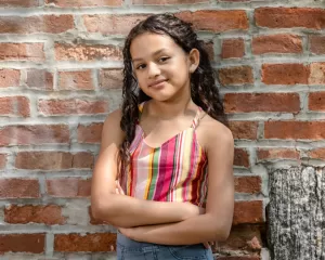 Professional Photographer A young girl with long curly hair stands in front of a brick wall, smiling softly with her arms crossed. She is wearing a colorful striped sleeveless top and jeans. Sunlight casts shadows, creating a natural, candid portrait perfect for headshots. www.maranonimagery.com