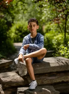 Professional Photographer A boy in a blue hoodie and shorts sits cross-legged on a large rock in a sunlit, green park, looking at the camera with a serious expression—an ideal setting for natural outdoor portraits. www.maranonimagery.com