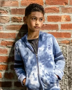 Professional Photographer A young boy wearing a blue tie-dye hoodie stands against a brick wall with his hands in his pockets, looking thoughtfully to the side in bright sunlight—an ideal subject for creative portraits by an aspiring photographer. www.maranonimagery.com