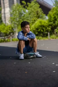 Professional Photographer A young boy sits on a skateboard with his arms resting on his knees, looking thoughtfully into the distance. Captured by a skilled photographer, he is outdoors on a paved path with trees and a bridge in the background. www.maranonimagery.com