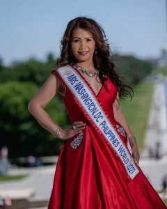 Professional Photographer A woman in a red dress and sash reading “Mrs. Washington DC Philippines World 2021” stands outdoors, smiling with one hand on her hip. Perfect for portraits, this photographer skillfully captures her against lush greenery and an out-of-focus walkway. www.maranonimagery.com