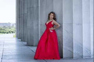 Professional Photographer A woman in a sleeveless red gown stands smiling and leaning against a marble column outdoors. Perfect for wedding photography, she wears jewelry, holds a small clutch, and is framed by the elegant stone architecture of a formal location. www.maranonimagery.com