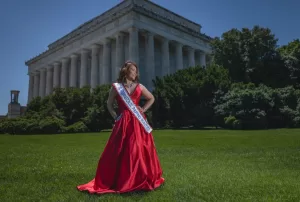 Professional Photographer A woman in a red gown and pageant sash stands on a grassy lawn in front of the Lincoln Memorial, posing confidently for the photographer on a sunny day, with trees and vibrant colors capturing the essence of gorgeous portraits. www.maranonimagery.com