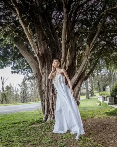 Professional Photographer A woman in a long, flowing white dress stands beneath a large, branching tree. She gazes thoughtfully into the distance, one hand touching her face—a serene moment perfect for wedding photography. Green grass and a road are visible in the background. www.maranonimagery.com