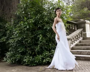 Professional Photographer A woman in a flowing white dress stands outdoors near stone steps and lush green bushes, looking to her left with one hand holding her dress—an elegant moment captured by a skilled wedding photographer. www.maranonimagery.com