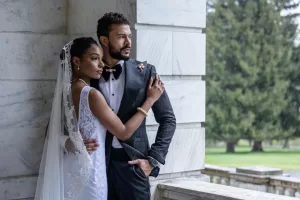 Professional Photographer A bride in a white lace dress and veil embraces a groom in a dark suit with a bow tie. They stand close together on a marble balcony, gazing into the distance, as their photographer captures timeless portraits with greenery in the background. www.maranonimagery.com