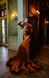 Professional Photographer A woman in an elegant, long, rust-orange velvet gown with a dramatic train poses for stunning wedding photography in a richly decorated hallway with vintage green and brown wooden walls and soft, warm lighting. www.maranonimagery.com
