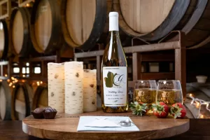 Professional Photographer A bottle of white wine, two filled glasses, candles, strawberries, and chocolates are arranged on a round wooden tray in front of wine barrels decorated with string lights—perfect for capturing romantic portraits. www.maranonimagery.com