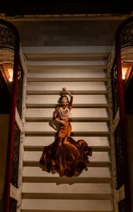 Professional Photographer A woman in an elegant, flowing brown dress with layered pearl necklaces poses dramatically on a grand staircase, perfect for striking portraits. Marble railings and warm lighting on either side complete this sophisticated scene. www.maranonimagery.com