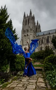 Professional Photographer A person in a vibrant blue dress poses gracefully on a stone path, flowing fabric extended like wings. Tall, gothic cathedral towers rise behind them under a cloudy sky—an inspiring scene for any photographer or lover of wedding photography. www.maranonimagery.com