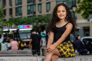 Professional Photographer A young girl with long curly hair, wearing a black tank top and yellow plaid skirt, sits on a stone ledge outdoors, smiling. Perfect for portraits, this lively scene includes blurred people and a colorful truck in the background. www.maranonimagery.com