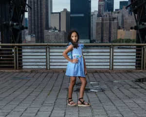 Professional Photographer A young girl in a blue dress and sandals poses confidently on a paved walkway beside a metal railing, with a city skyline and river in the background—perfect for creative portraits. www.maranonimagery.com