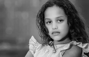 Professional Photographer Black and white photo of a young girl with curly hair, wearing a striped ruffled top and a necklace, looking thoughtfully at the camera—a striking headshot that captures her neutral expression. www.maranonimagery.com