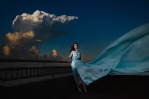 Professional Photographer A woman in a flowing light blue dress stands on a balcony at dusk, her gown billowing dramatically in the wind—a scene reminiscent of elegant wedding photography—set against dark sky and large, illuminated clouds. www.maranonimagery.com