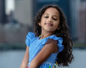 Professional Photographer A young girl with long curly hair wearing a blue dress with ruffled sleeves poses confidently outdoors, captured beautifully by a talented portrait photographer against a blurred cityscape background. www.maranonimagery.com