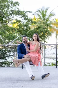 Professional Photographer A man in a blue suit and a woman in a coral dress sit together on a hanging net swing outdoors, smiling and holding each other, with palm trees and water in the background—capturing a perfect moment for portraits by a skilled photographer. www.maranonimagery.com