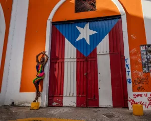 Professional Photographer A person in a colorful dress poses against a wall painted with the Puerto Rican flag on large doors, creating a vibrant backdrop perfect for creative headshots. The orange wall and arch are covered in graffiti and stickers on one side. www.maranonimagery.com