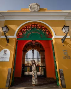 Professional Photographer A woman in a floral dress stands in front of the bright yellow entrance arch of the Museo de San Juan, as a photographer captures portraits framed by green and red details above the gate and two black lanterns on either side. www.maranonimagery.com