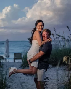 Professional Photographer A man lifts a smiling woman in a white dress at a sandy beach with grass, lounge chairs, and ocean waves in the background under a cloudy sky, creating a scene as timeless as classic portraits. www.maranonimagery.com