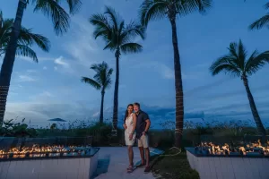 Professional Photographer A couple stands close together on a path lined with palm trees and glass fire pits at sunset, as a photographer captures their smiles against the blue sky and clouds. They are dressed in summer clothing, radiating warmth and joy. www.maranonimagery.com