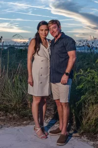 Professional Photographer A woman in a light-colored dress and a man in a dark shirt and shorts stand together, smiling, on a sandy path with grasses and a sunset sky in the background—an ideal moment captured by an expert portrait photographer. www.maranonimagery.com