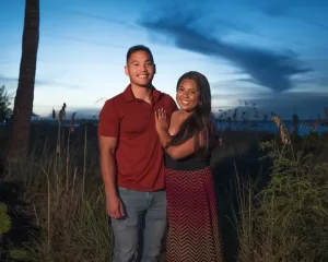 Professional Photographer A smiling couple stands close together outdoors at sunset, with tall grass and a palm tree behind them. Captured by a talented photographer, the man wears a red shirt and jeans; the woman wears a patterned skirt and dark top under the blue, cloud-speckled sky. www.maranonimagery.com
