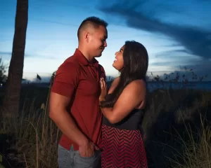 Professional Photographer A couple stands close together outdoors at dusk, smiling at each other. Captured by a skilled wedding photographer, they pose amid tall grass and a darkening blue sky—the man in a red shirt, the woman in a black and red dress. www.maranonimagery.com