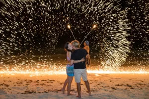 Professional Photographer A couple kisses on a sandy beach at night while a performer behind them spins flaming sticks, creating a dramatic shower of sparks—captured perfectly by a photographer known for stunning portraits and creative pet photography. www.maranonimagery.com