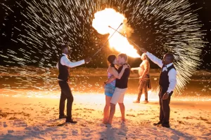 Professional Photographer A couple kisses on a beach at night while two performers, dressed in vests and shirts, create a fiery display with torches. Captured by a talented wedding photographer, sparks dance in the air behind them as another performer stands holding fire. www.maranonimagery.com