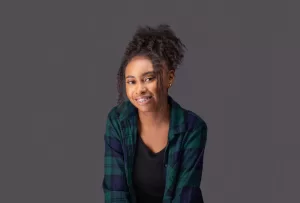 Professional Photographer A young woman with curly hair and braces smiles slightly, wearing a green and blue plaid shirt over a black top, posing for photographer-style headshots in front of a plain gray background. www.maranonimagery.com