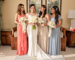 Professional Photographer A bride in a white dress stands indoors with three bridesmaids in coral, beige, and blue gowns. This candid portrait by a skilled photographer captures them smiling with bouquets in a warmly lit room adorned with wooden furniture and floral artwork. www.maranonimagery.com
