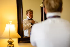 Professional Photographer A man stands in front of a mirror, adjusting his necktie while wearing a white shirt. A photographer’s camera rests nearby on the table beside a lamp with a beige shade, and the warmly lit room exudes quiet anticipation. www.maranonimagery.com