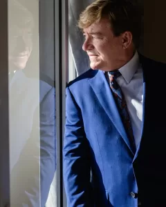Professional Photographer A man in a blue suit and patterned tie stands by a window, looking outside with sunlight on his face. His reflection is visible in the glass, capturing the poised look of a professional photographer ready for headshots. www.maranonimagery.com