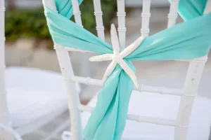 Professional Photographer A white chair decorated with a turquoise fabric drape, tied in the center with a white starfish ornament, suggesting a beach or ocean-themed wedding—perfect for adding charm to your wedding photography. www.maranonimagery.com